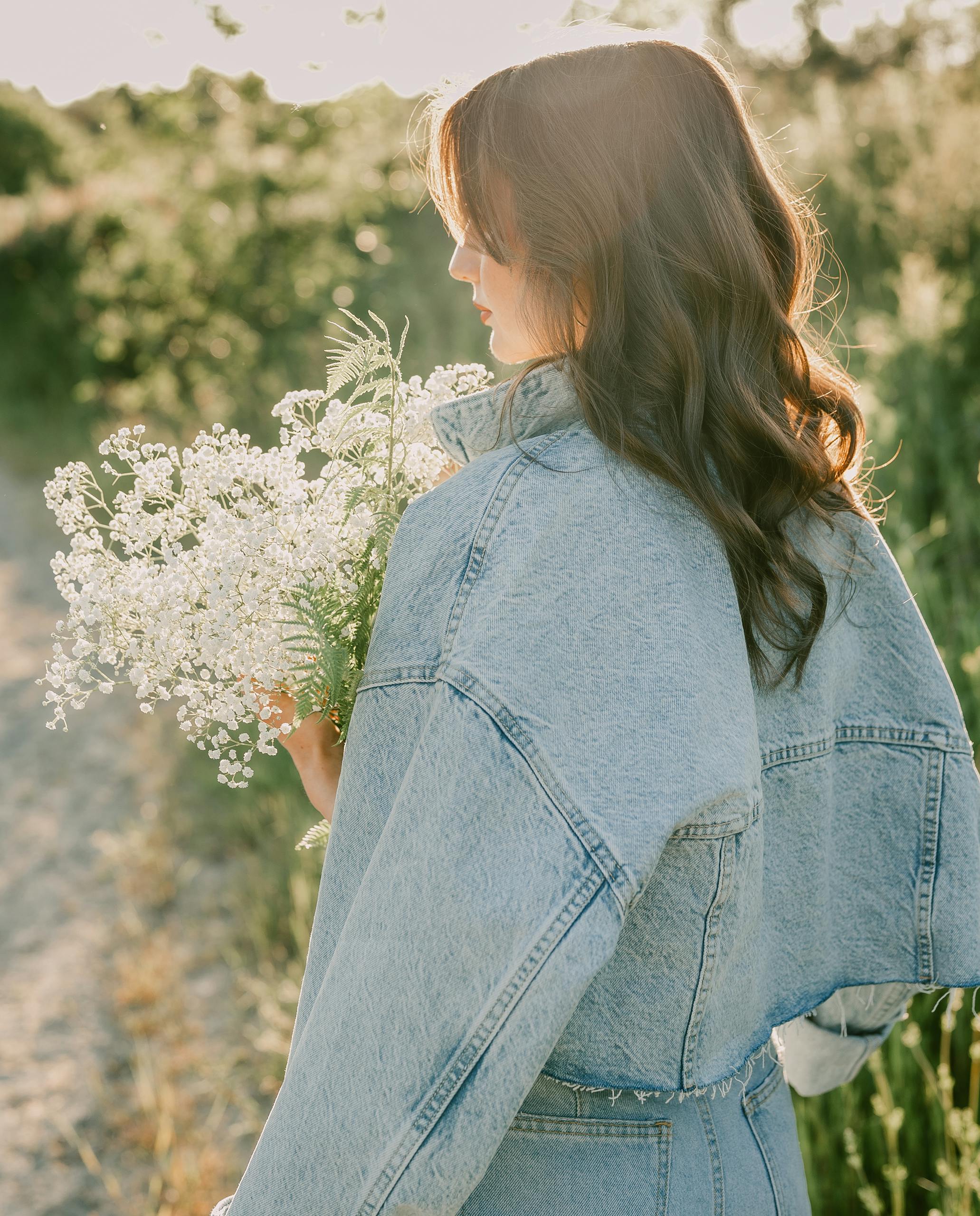 Brunette woman in a denim jacket holding delicate flowers on a sunny day outdoors.