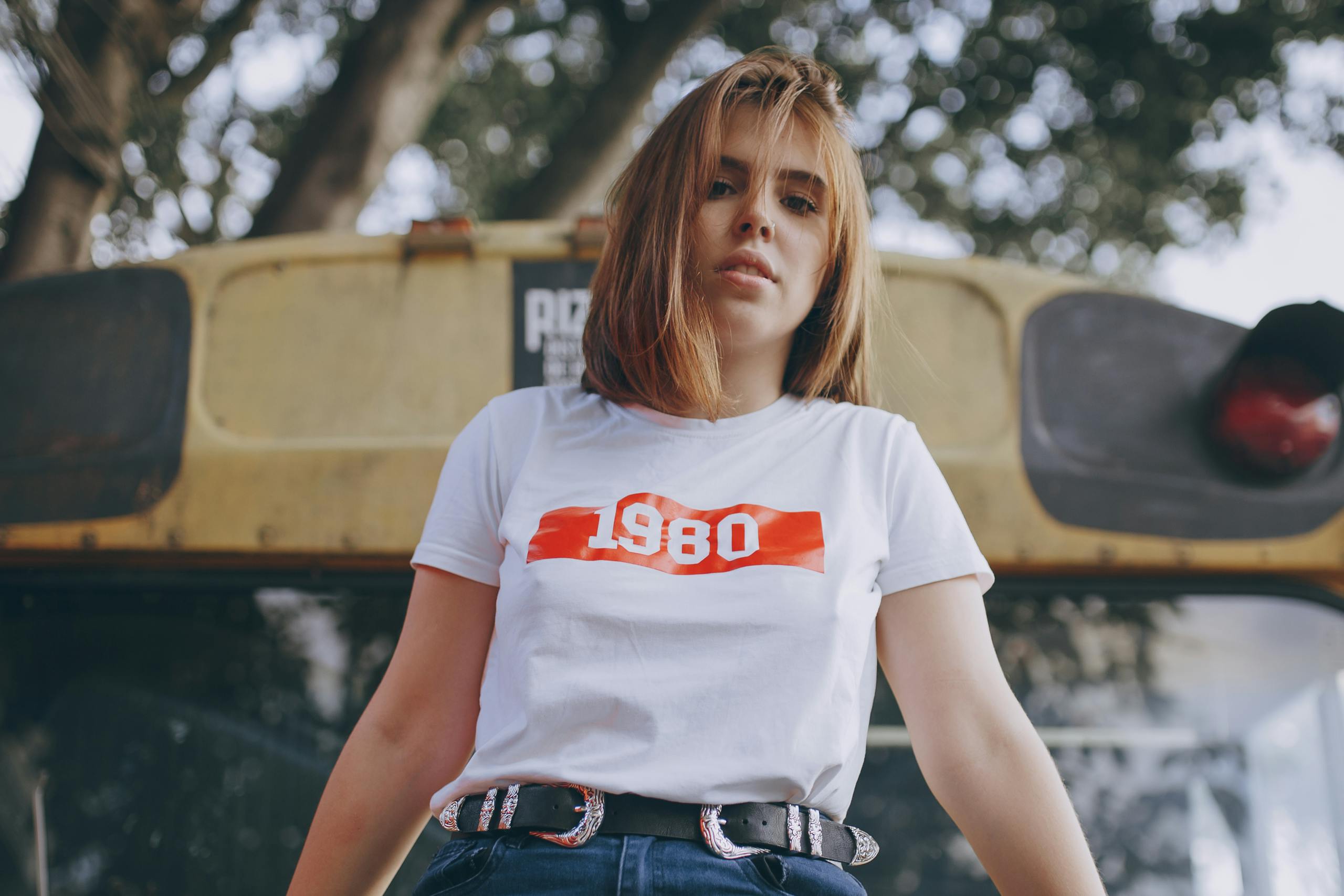Stylish young woman in a 1980 shirt poses confidently in front of a vintage bus outdoors.