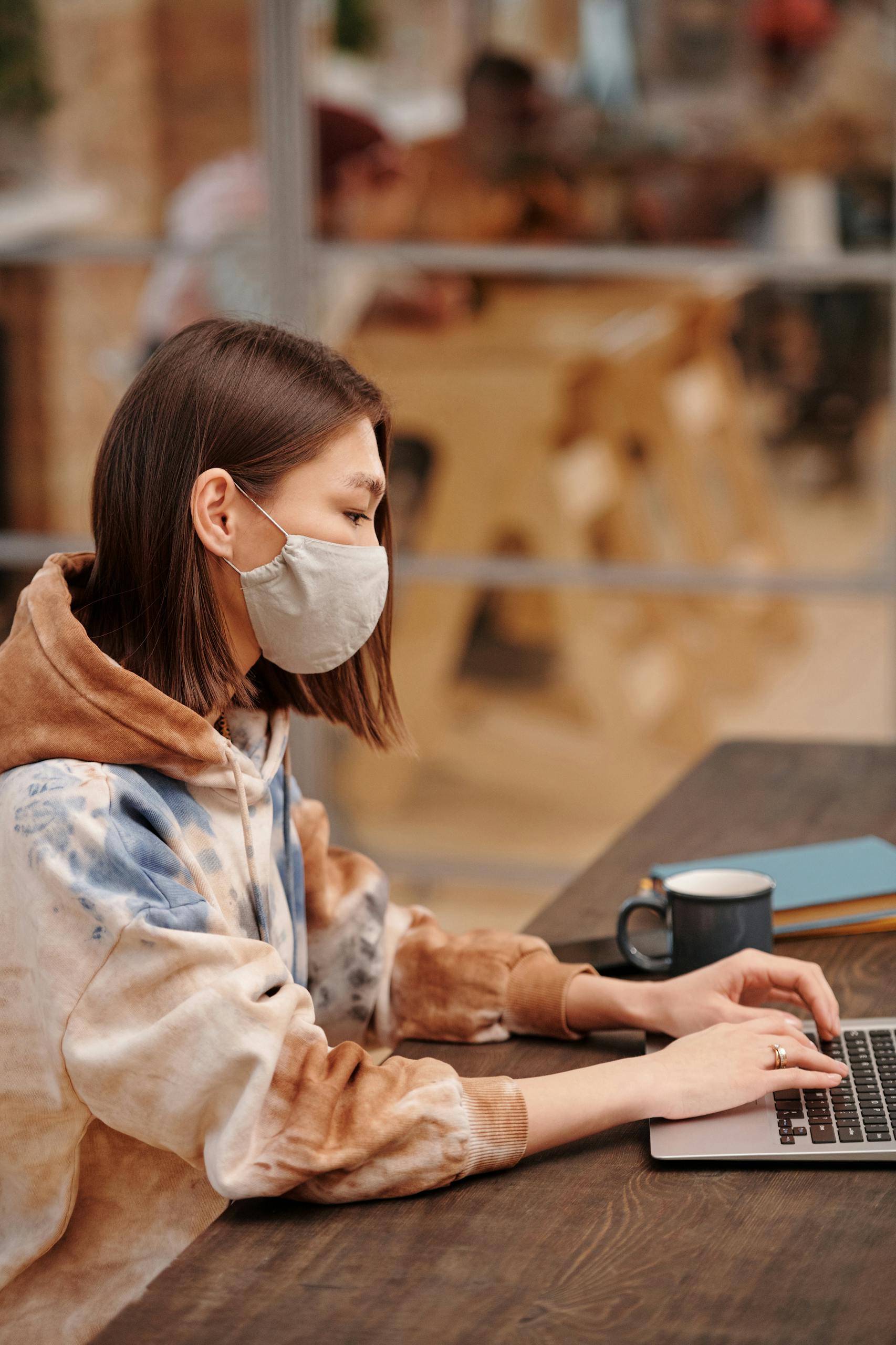 Woman in facemask typing on a laptop at a wooden table, emphasizing modern technology and remote work.